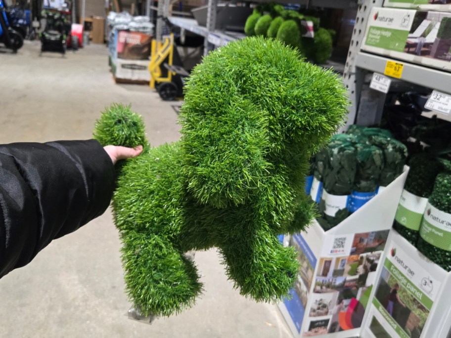 A hand holds a decorative artificial grass sculpture shaped like a dog inside a hardware store. Shelves with garden supplies are in the background.