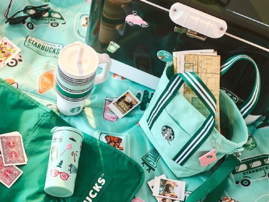 Stylish scene with Starbucks merchandise: mint green bag, map, colorful tumblers, polaroid photos, playing cards, and sunglasses on a playful mat.