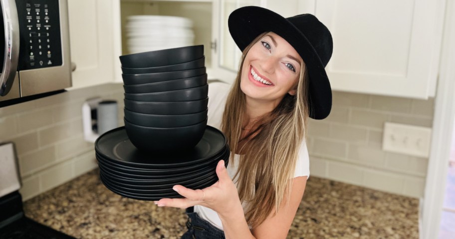A woman in a black hat, smiling while holding a stack of black dishes and bowls in a kitchen with light cabinets and tiled backsplash.