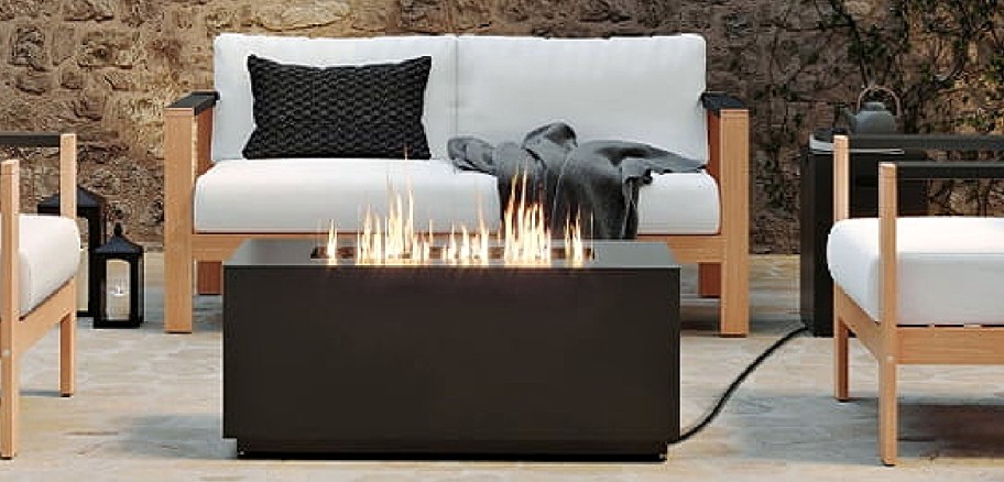 A cozy outdoor patio setting features a modern fire pit table with a warm flame, surrounded by cushioned wooden chairs.