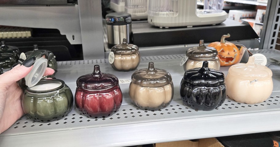 small glass pumpkin jar candles in a row on store shelf with hand taking the lid off one of them