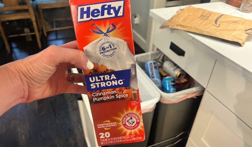Person holding box of hefty ultra, strong, cinnamon, spice, trash bags, next to kitchen, trashcan, and Amazon packaging