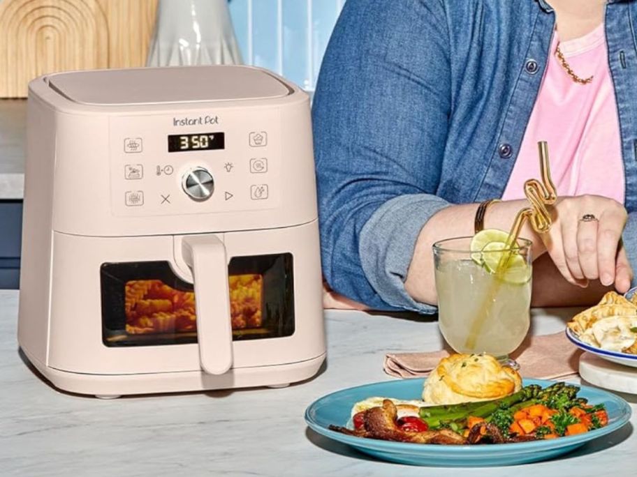 An instant Pot Mini Air Fryer next to a woman with a plate of food and a beverage in a glass