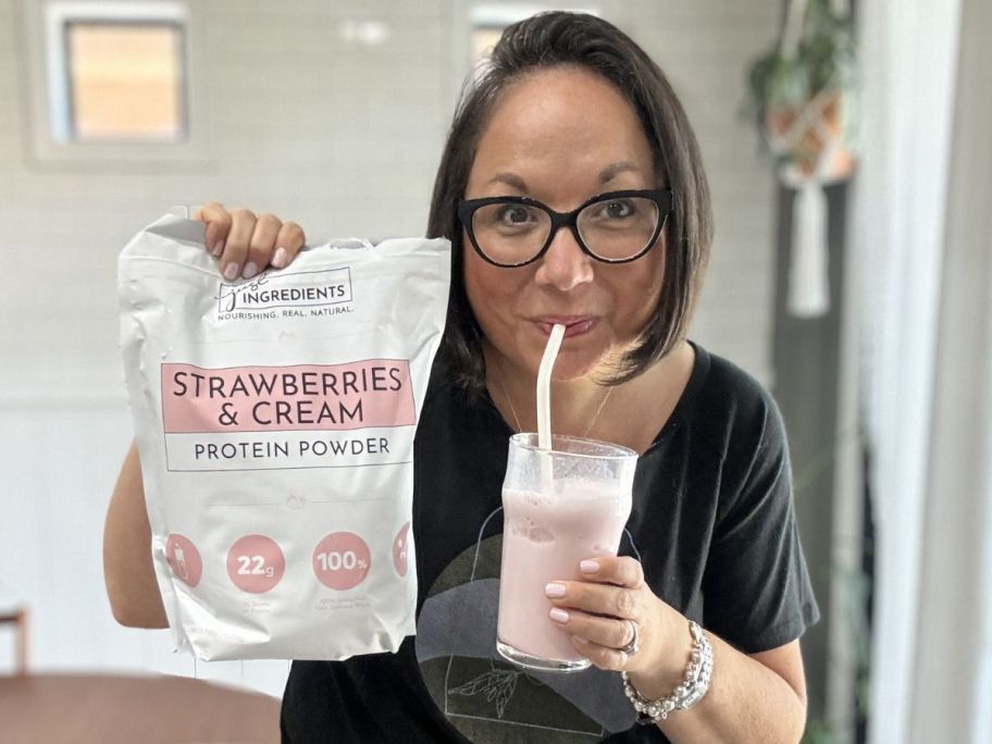 Woman drinking a pink protein shake while holding a big bag of Just Ingredients Protein Powder
