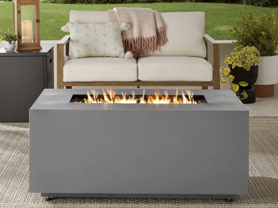 Modern outdoor patio with a sleek gray fire pit table, vibrant flames flickering. Beige loveseat with cozy pillows and a pink throw blanket in background.