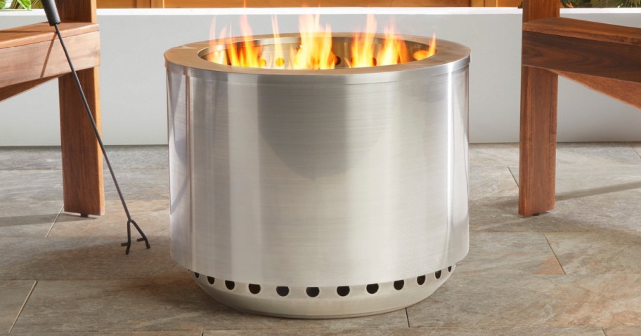 A sleek stainless steel fire pit with visible flames, surrounded by wooden furniture and a stone patio.