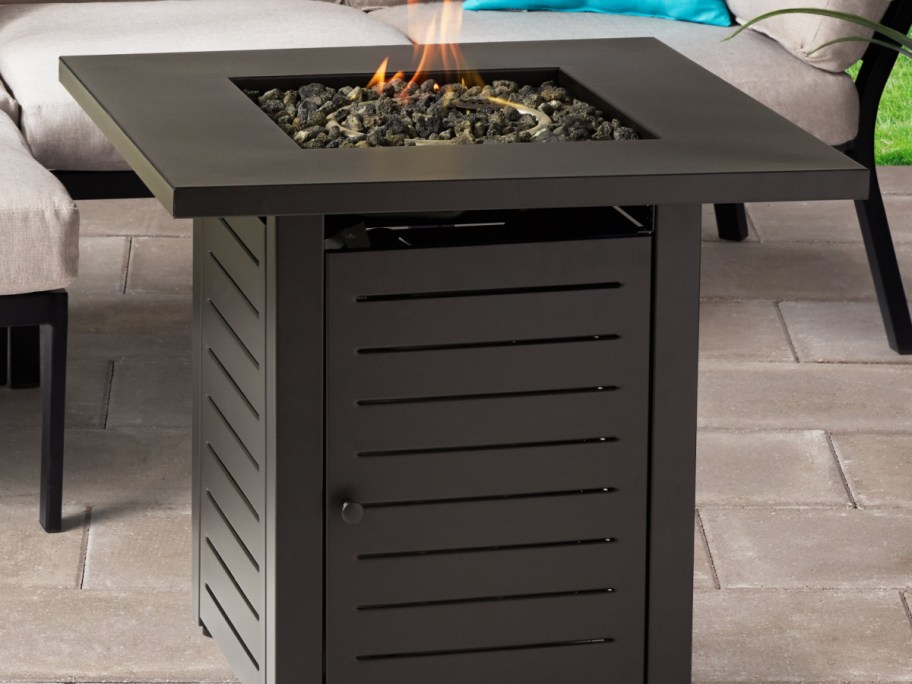 A sleek, square fire pit table with a metal base and open flame on top. It rests on a patio, surrounded by plush outdoor furniture.