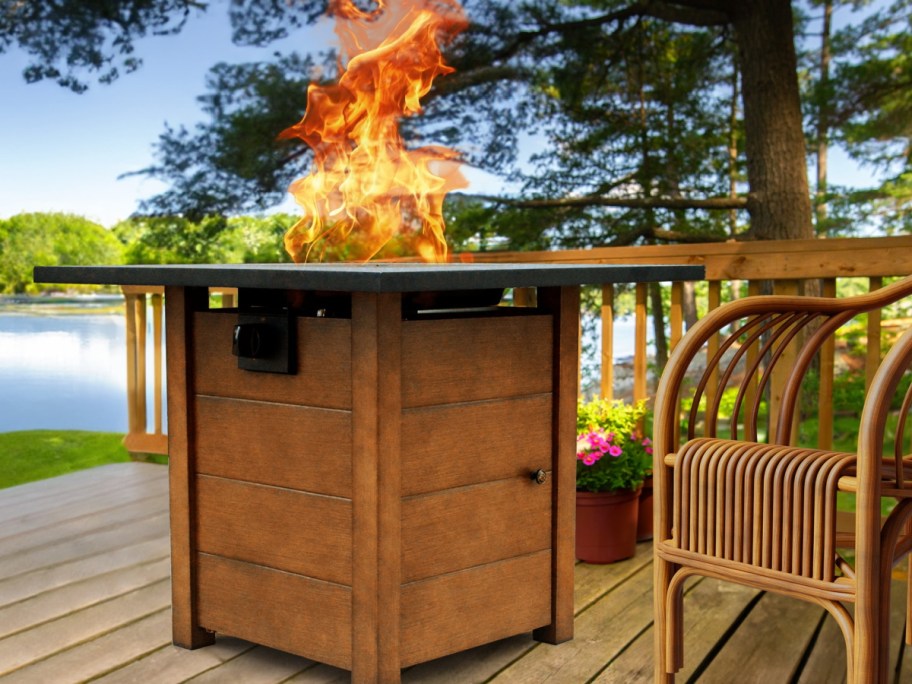Fire pit on a wooden deck by a lake, with flames rising high. Surrounded by trees and a wicker chair.