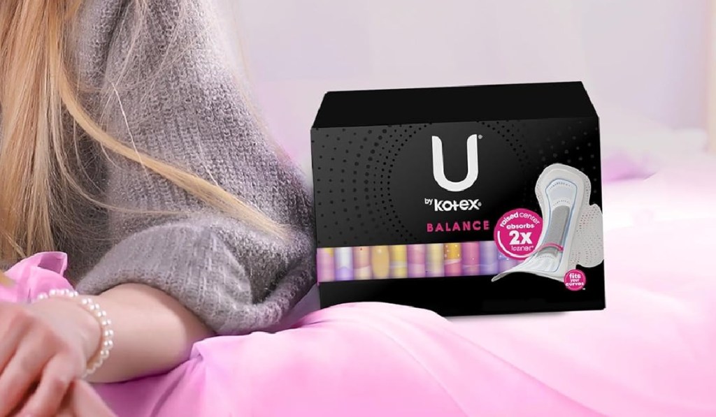 Box of Kotex pads next to women wearing gray sweater and sitting on pink comforter