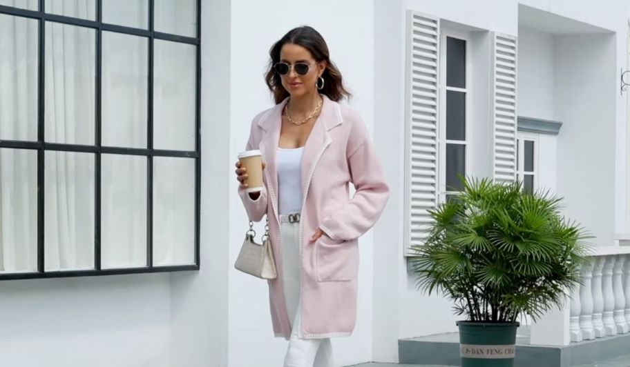 a woman in a pink coatigan walking down the sidewalk with a cup of coffee