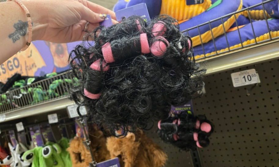 a womans hand holding a black curly dog wig with pink foam rollers