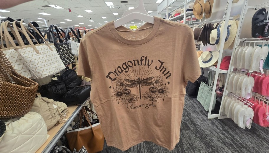 a woman holding a tan graphic tee by the hanger