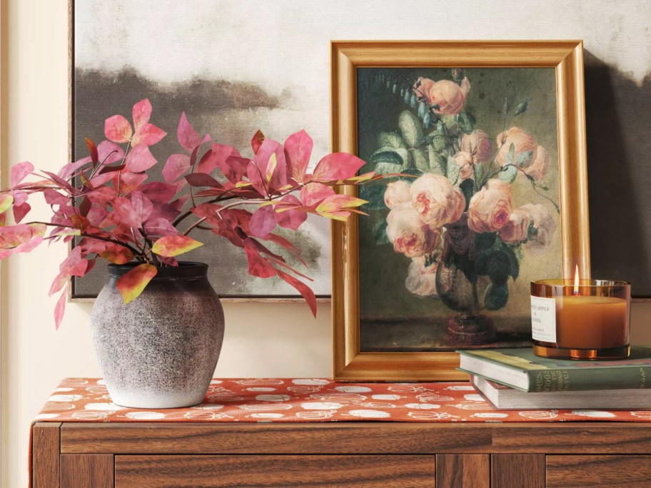 A cozy scene featuring a vase with pink leaves, a framed floral painting, a lit candle, and stacked books on a wooden table with a patterned cloth.