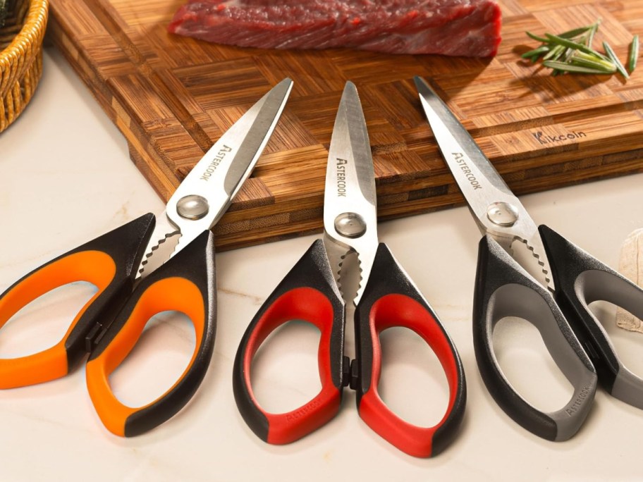 Three pairs of multi-colored kitchen shears with orange, red, and gray handles are placed on a counter near a wooden cutting board with raw meat.