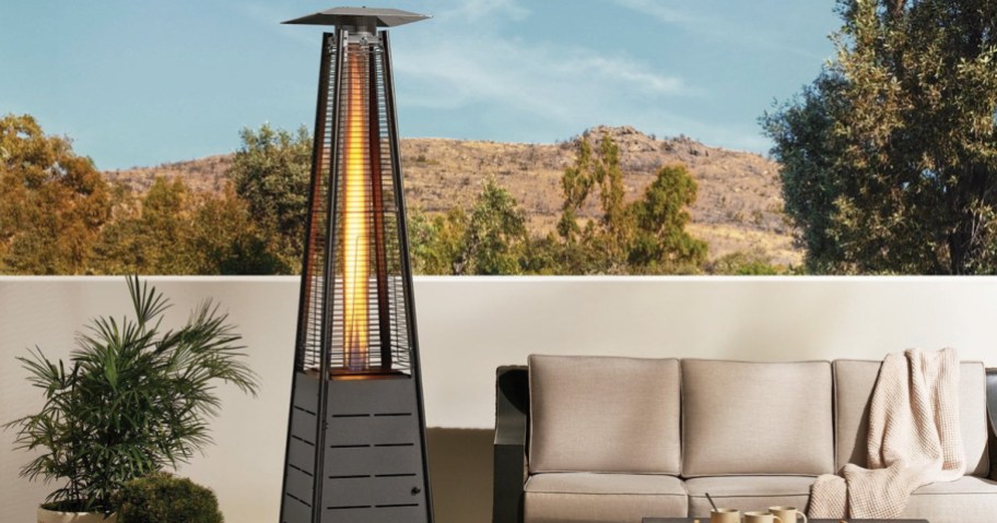 Outdoor patio with a tall, lit patio heater next to a beige sofa adorned with a blanket. Mountain view and trees in the background under a clear sky.