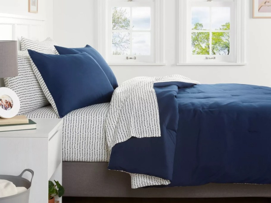 A neatly made bed with a navy blue comforter and patterned sheets is set in a bright room. Two windows show a view of trees outside.