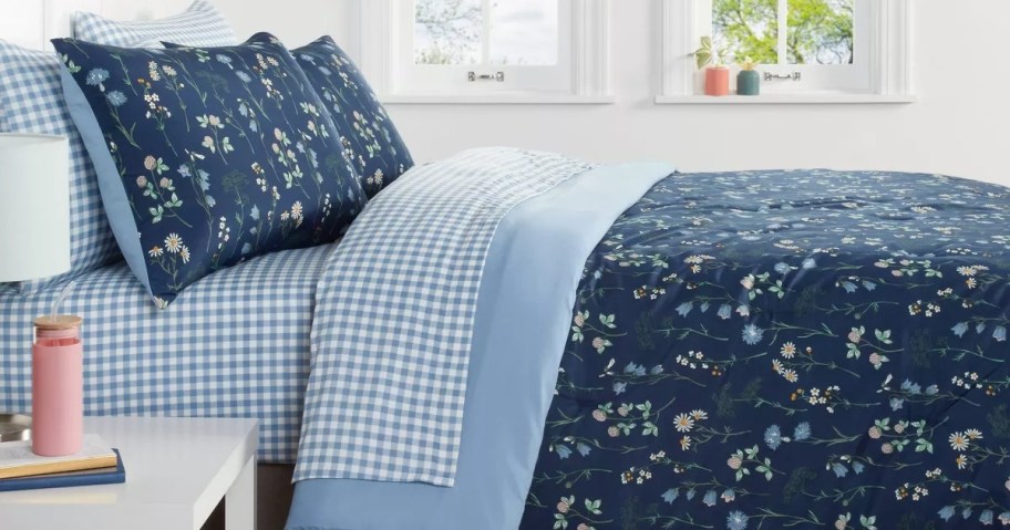 A cozy bed features navy floral-patterned bedding and blue gingham sheets, next to a white nightstand with books and a pink thermos, under a sunny window.