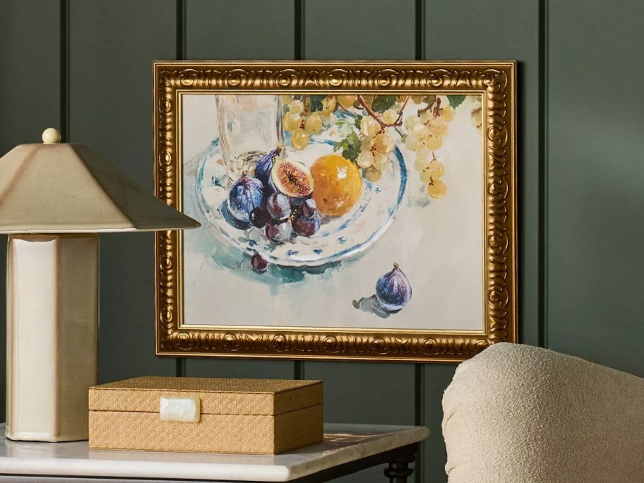 A cozy corner with a beige armchair and throw pillow, a marble-top side table holding a lamp, framed fruit painting on a green paneled wall.