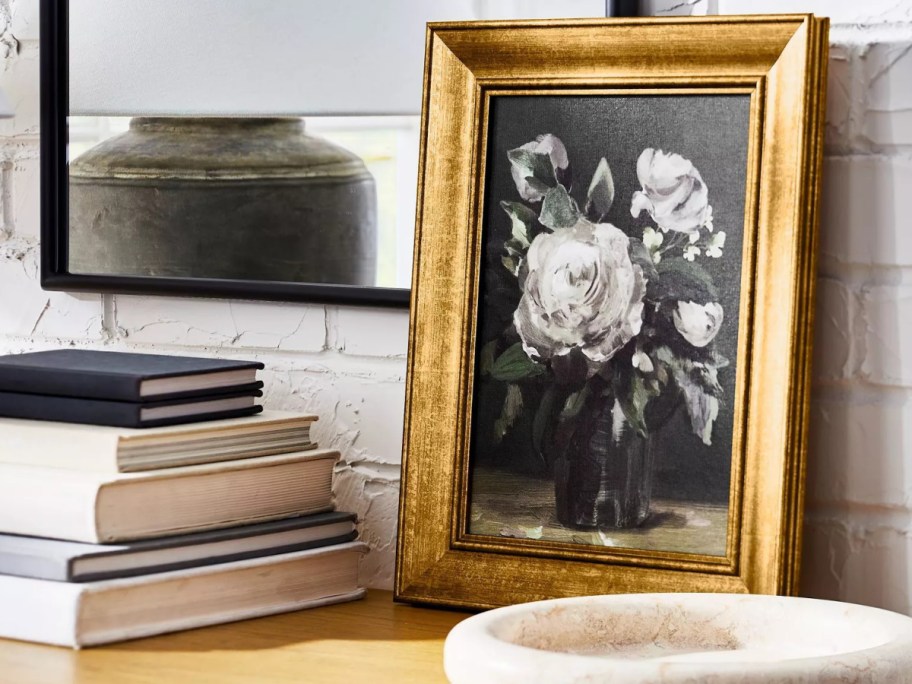 A gold-framed floral painting of white roses on a wooden shelf next to a stack of books, with a mirror reflecting a vase against a white brick wall.