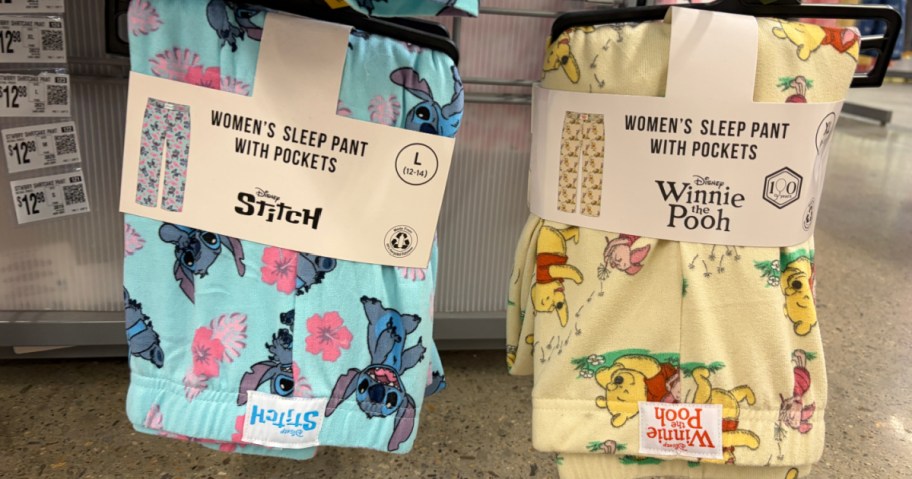 Two pairs of women's sleep pants, one featuring a blue design with Stitch and flowers, and the other a yellow design with Winnie the Pooh characters. Both have pockets and are displayed on hangers in a store.