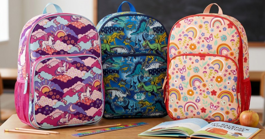Three colorful backpacks on a classroom desk: one with unicorns and clouds, another with dinosaurs, and a third with rainbows and flowers. Pencils, a ruler, and an open book are nearby, along with an apple.