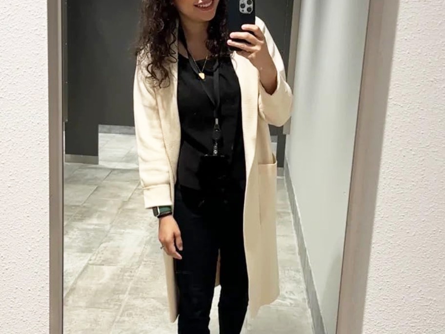woman taking photo in a mirror wearing a black outfit with a white coat
