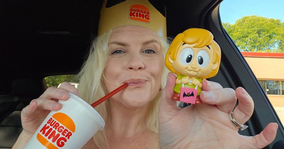 A blonde woman holding a small plastic toy while wearing a crown and sipping on a soda with a straw