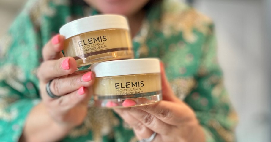 A person wearing a green patterned shirt holds two jars of Elemis Pro-Collagen Cleansing Balm, with pink nail polish contrasting the soft-focus background.