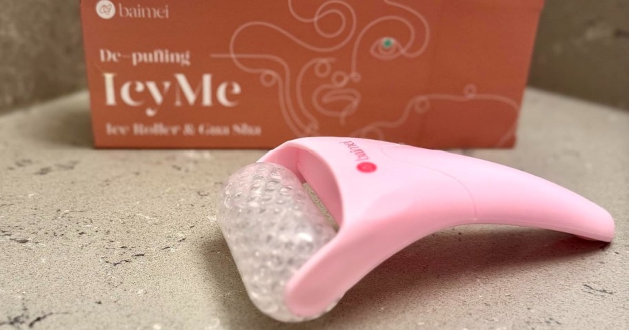 Pink icy roller with textured surface on gray countertop in focus. Background shows an orange box with abstract line art and the words "De-puffing IcyMe."