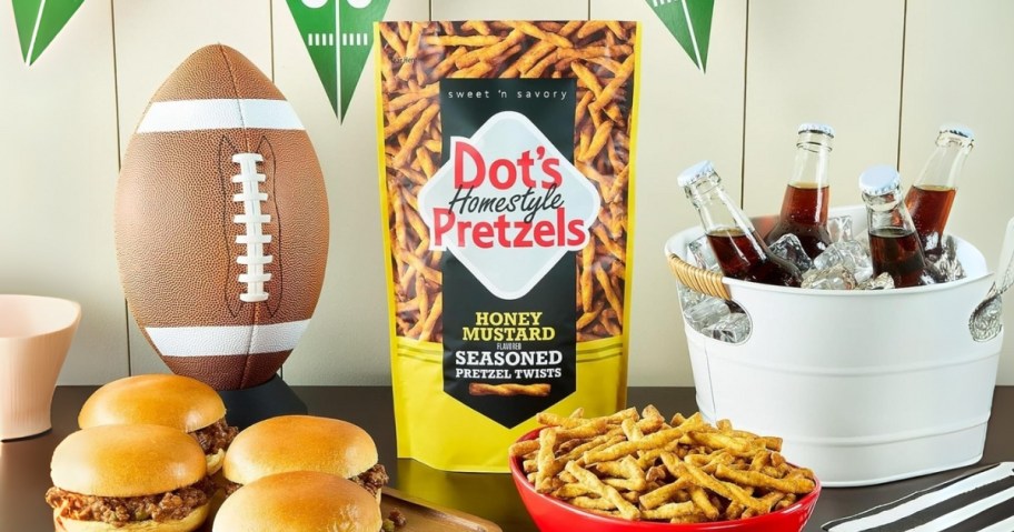 a bag of Dots Honey Mustard Pretzels on a table with football decor, a tub of drinks, burgers, and a bowl of the pretzels