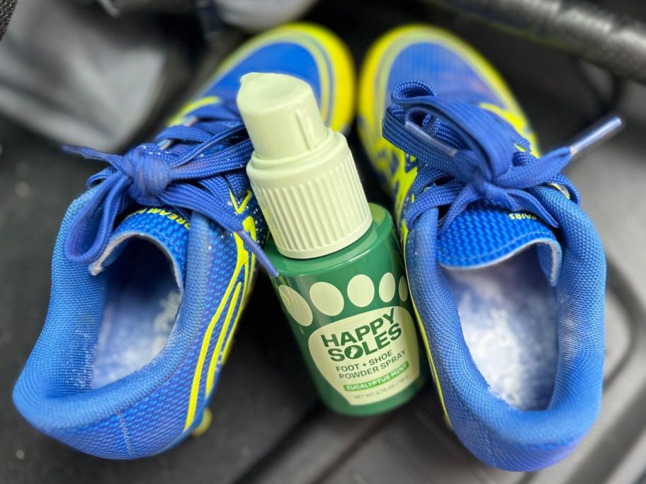 A bottle of Happy Soles Foot & Shoe Deodorizer Powder Spray in between two kids cleats