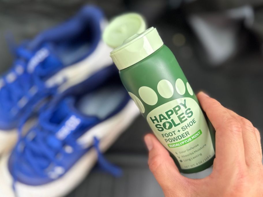 Hand holding a bottle of Happy Soles Shoe Deodorizer & Foot Powder over a pair of cleats