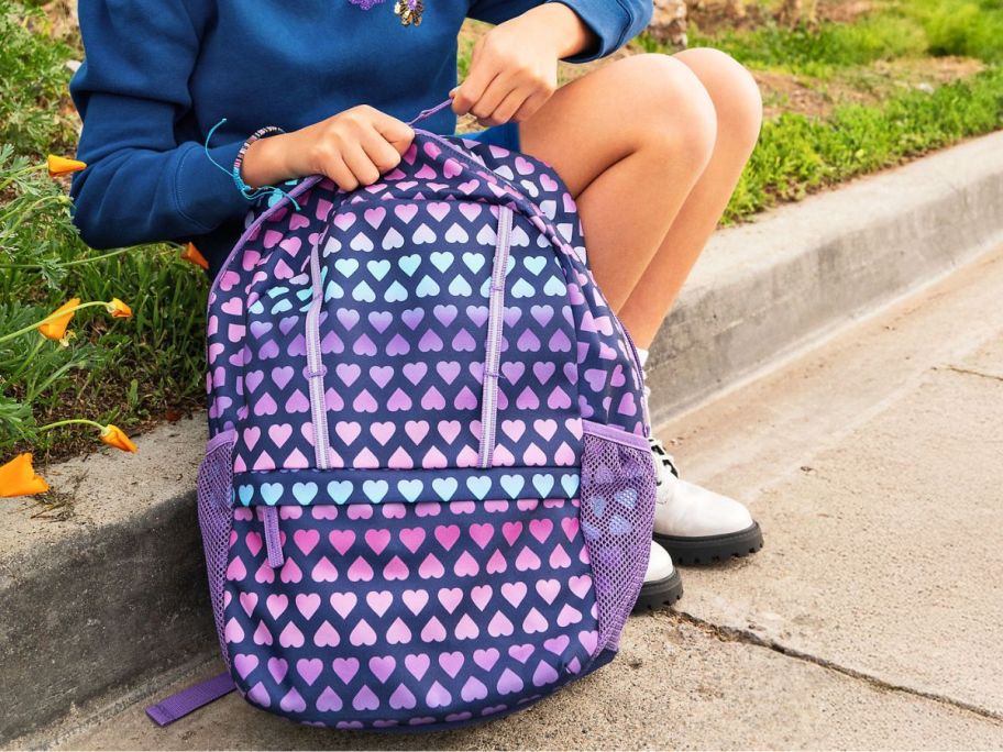 A child sitting on a curb with a heart-themed backpack