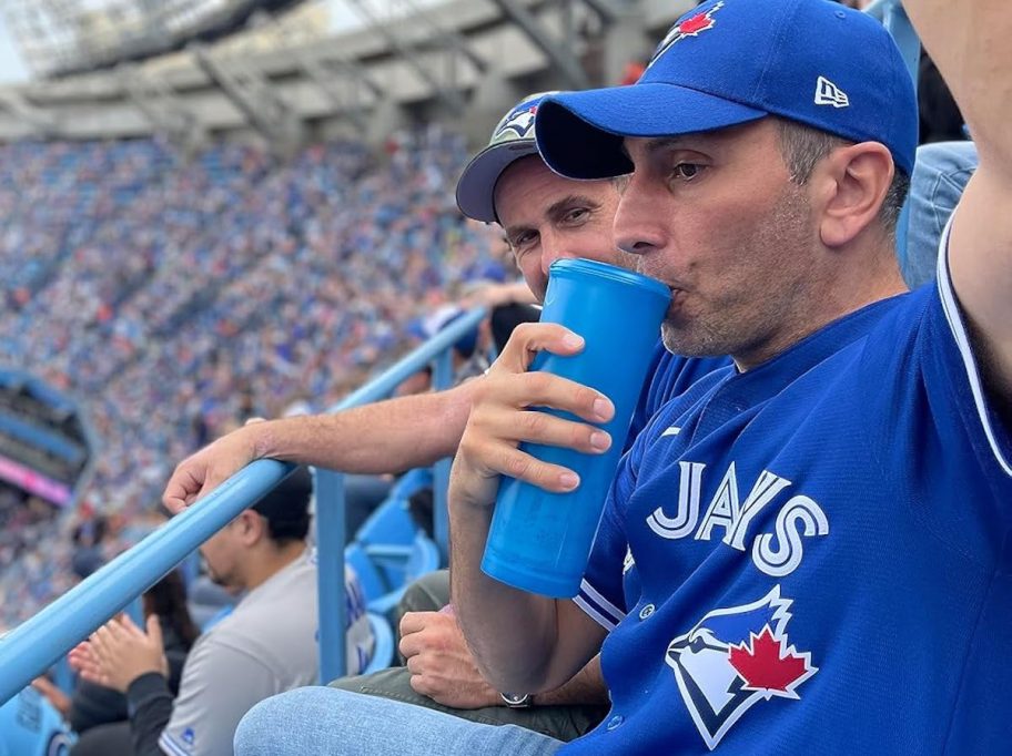 Man in blue Jays sports gear sitting in stadium sipping from blue tumbler 