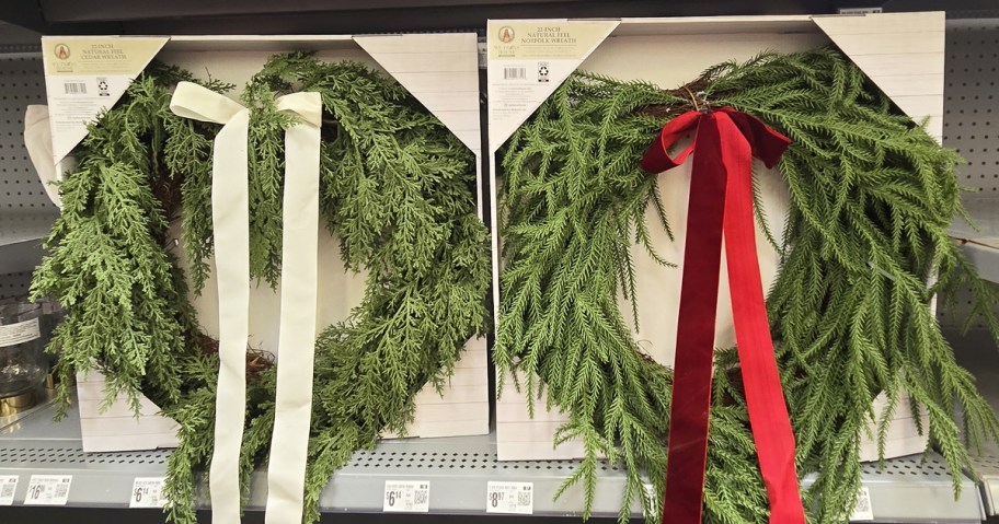 two faux christmas wreaths on store shelf, one with a white bow and one with a red bow