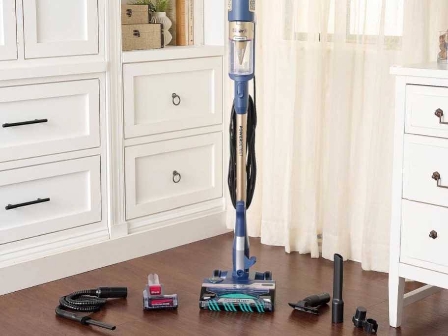 A sleek blue and gold vacuum cleaner with various attachments is arranged on a wooden floor in a bright room with white cabinets and sheer curtains.