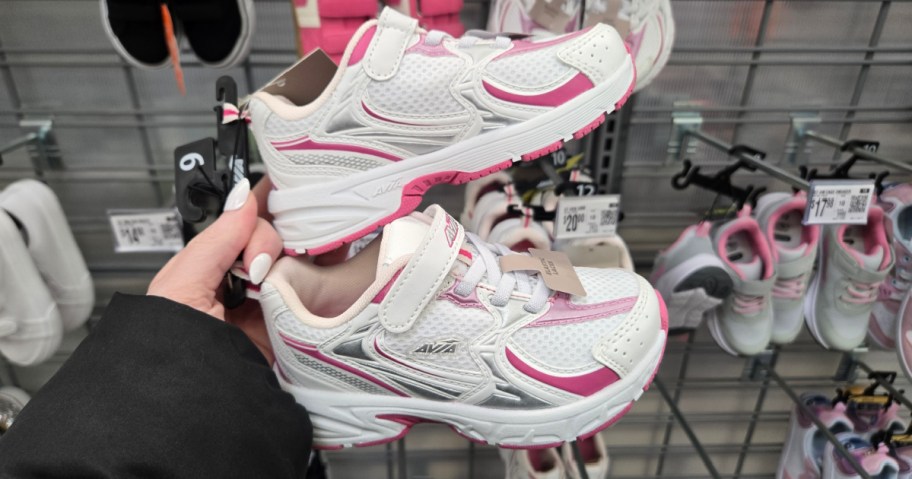 A hand holds up a pair of small white and pink children's sneakers with Velcro straps inside a store. Other colorful shoes are displayed in the background.