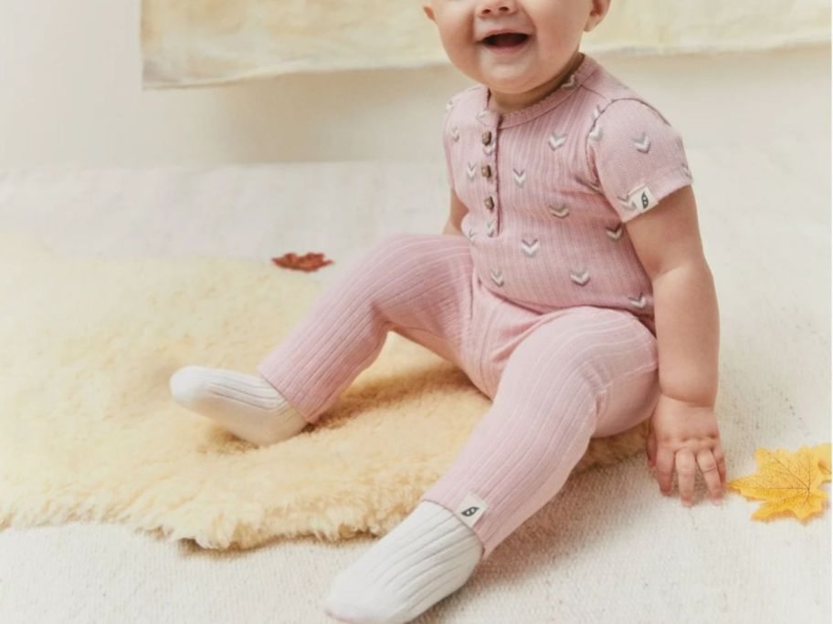 A baby girl sitting on the floor smiling wearing a pink short sleeved shirt and pants