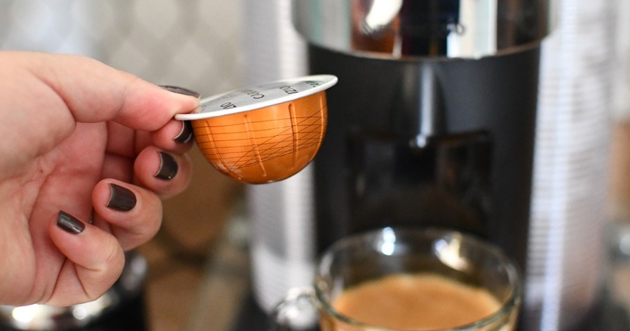 A hand with dark nails holds a coffee pod near a coffee machine, with a glass cup of espresso below.