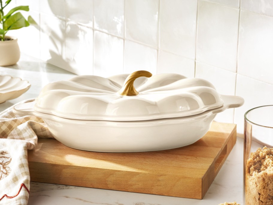 A large white ceramic pumpkin-shaped baking dish with a gold stem handle on its lid, sitting on a wooden cutting board.