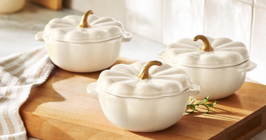 Three white, pumpkin-shaped ceramic dishes with gold stem handles on their lids sitting on a wooden cutting board.