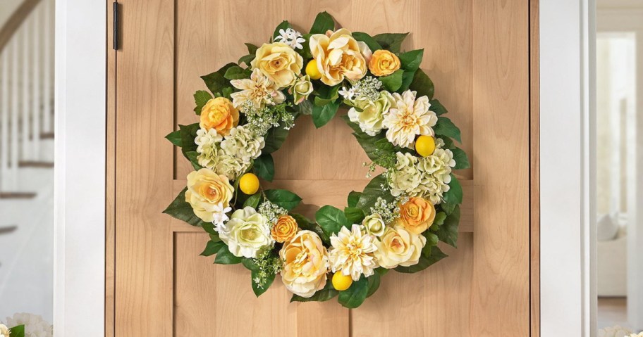 A floral wreath made of yellow and white flowers, green leaves, and small decorative lemons, hung on a wooden door.