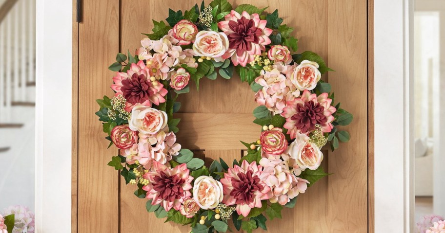 A floral wreath made of various pink and cream flowers, including roses and dahlias, displayed on a wooden door.