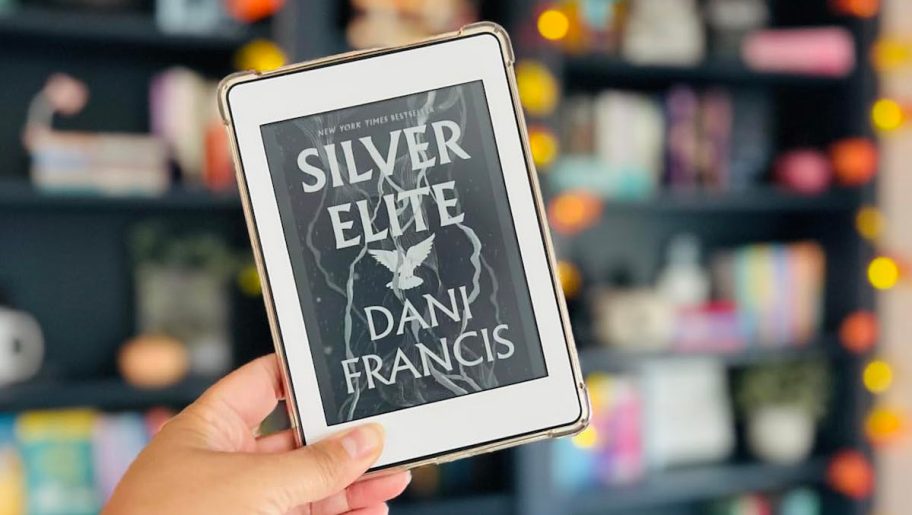 hand holding silver elite book on kindle in front of bookshelf