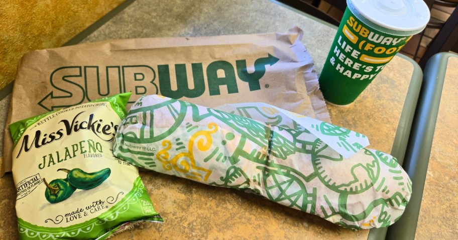 a Subway sub in the wrapper next to a bag of chips and a drink on a table