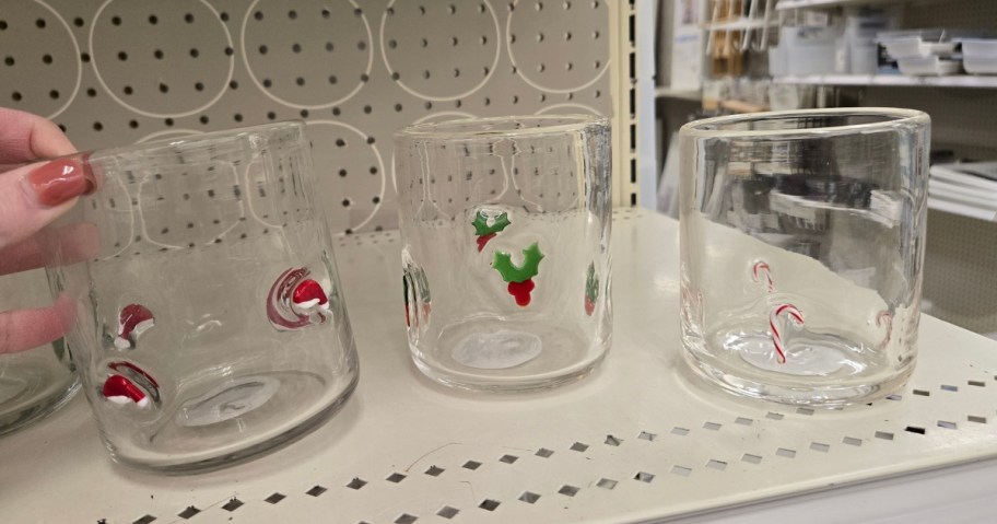 A hand reaching for a juice glass with a Santa hat on, there are more glasses with holly berries and candy canes next to it on the store shelf