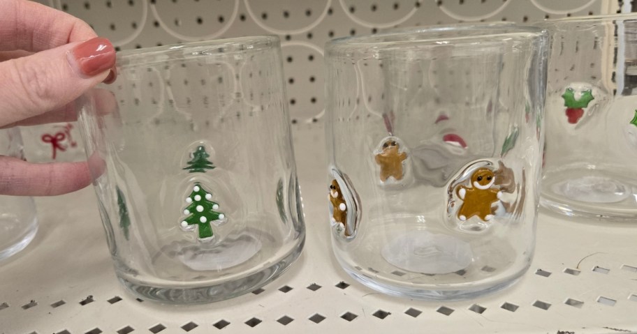hand reaching for a juice glass with Christmas trees on it next to a glass with Gingerbread men on a store shelf