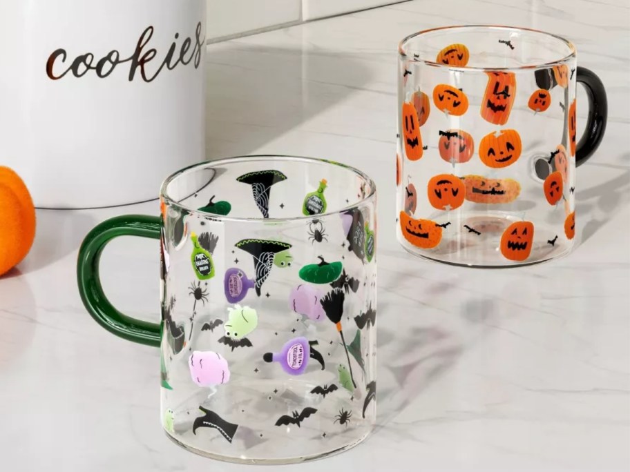 two glass coffee mugs with Halloween designs, one is witch hats and potions, the other is jack-o-lanterns