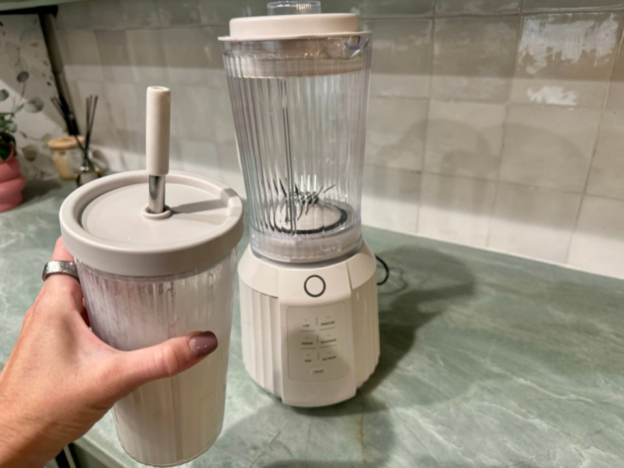 hand holding tumbler with straw and smoothie inside next to blender