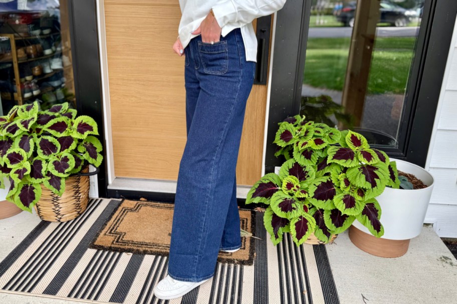 woman in dark wide leg jeans in front of door between two green leafy planters